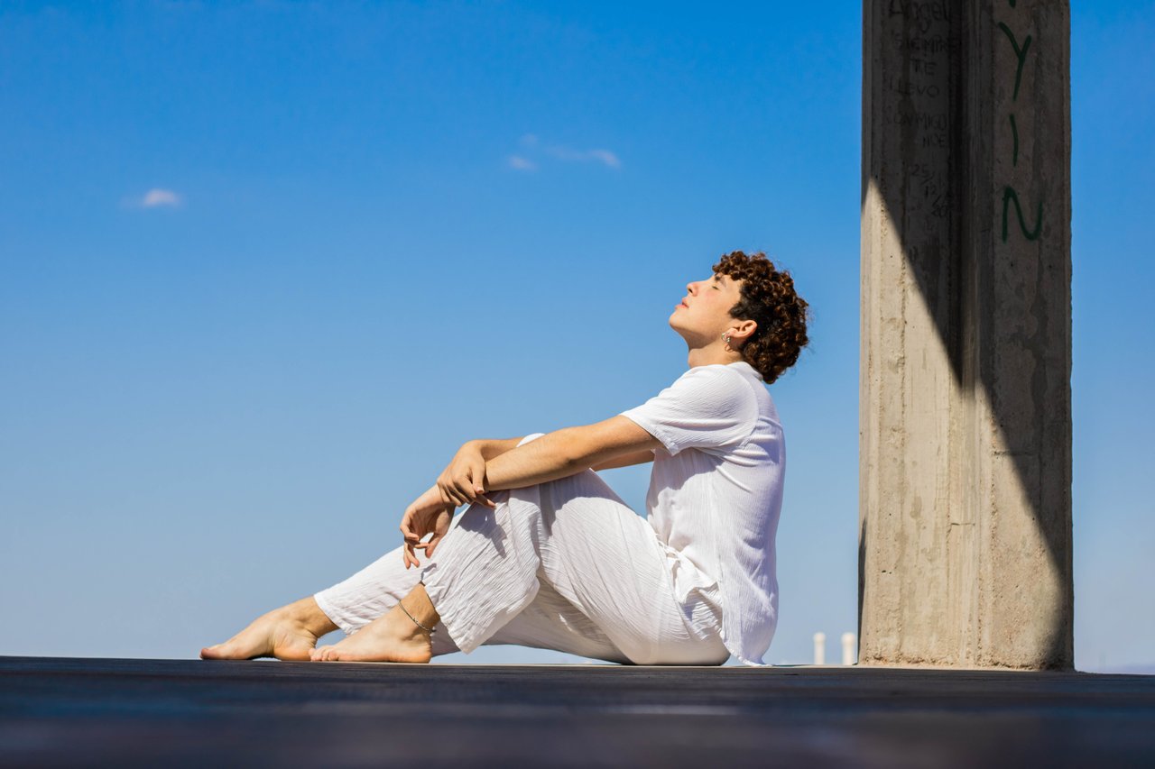 A man in white clothing sitting peacefully outdoors against a vibrant blue sky.