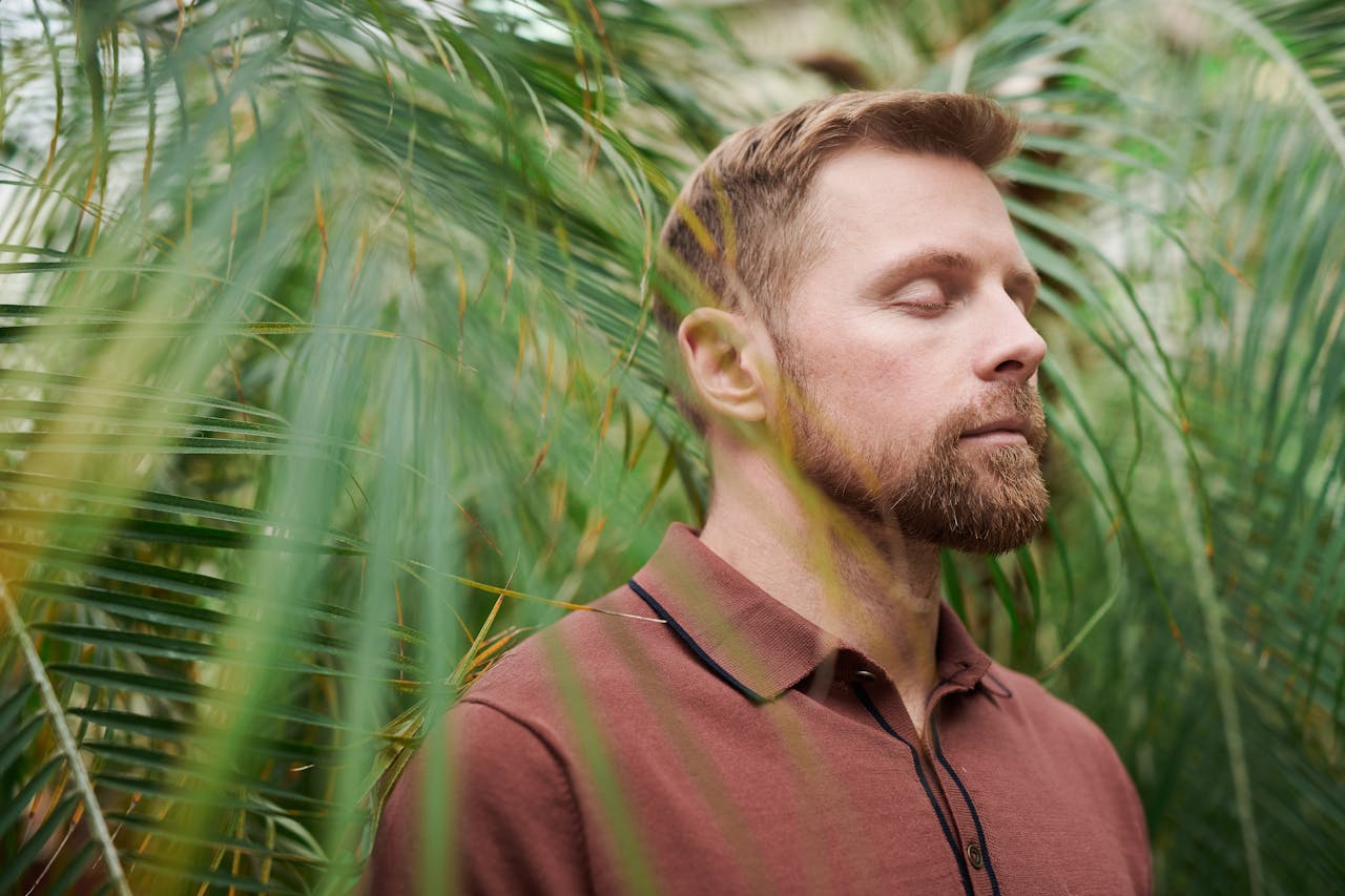 A man with closed eyes meditates peacefully amidst lush green palm leaves, promoting relaxation and mindfulness.